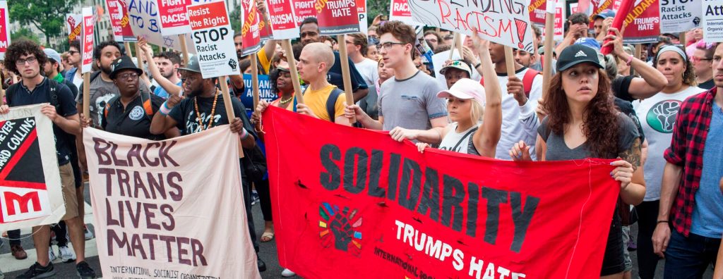 The image shows a crowd holding banners and placards. One large banner says, "Solidarity Trumps Hate," and another says, "Black Trans Lives Matter."