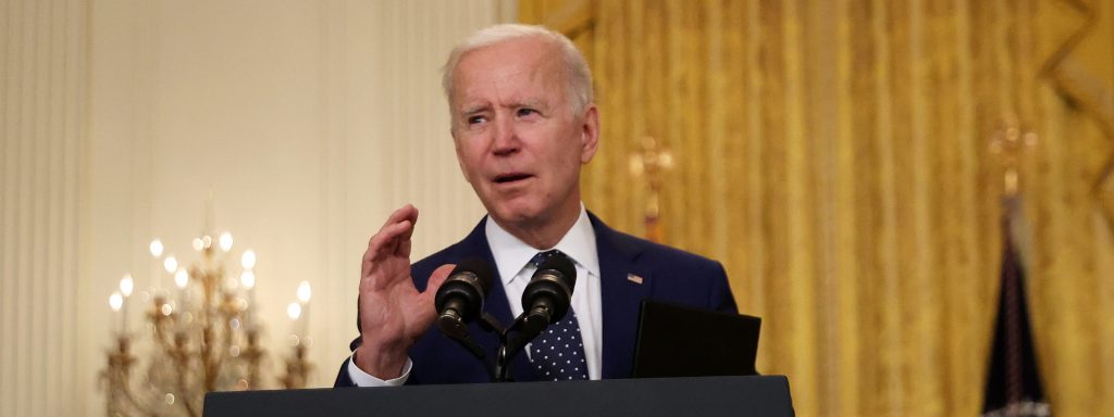U.S. President Joe Biden stands at a podium and announces new economic sanctions against the Russia government from the East Room of the White House on April 15, 2021 in Washington, DC. A chandelier-type lamp and multiple flags stand behind him.