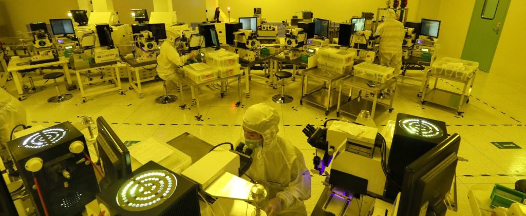 Employees dressed in white sanitary gowns, head coverings and surgical masks sit at stations in a yellow-hued room, making chips at a factory of Jiejie Semiconductor Company in Nantong, in eastern China's Jiangsu province on March 17, 2021. (Photo by STR/AFP via Getty Images)