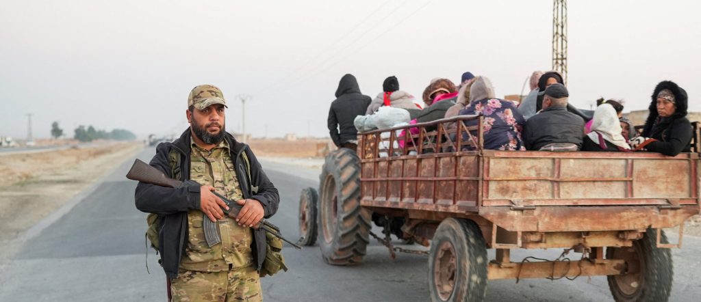 On the left, a man in a camo outfit stands in the street with a gun as an open-bed truck with people inside it drives away on the right.