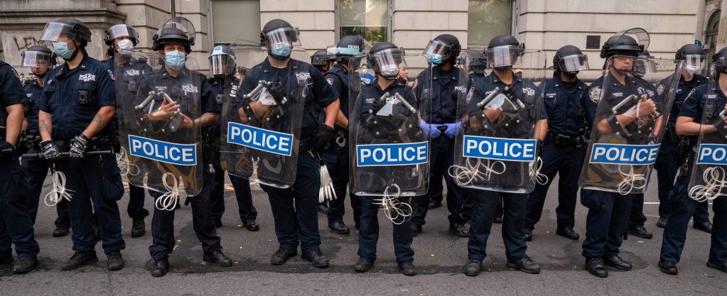 Police in full riot gear, some wearing masks and others not, stand in a row on July 1, 2020 in New York City.