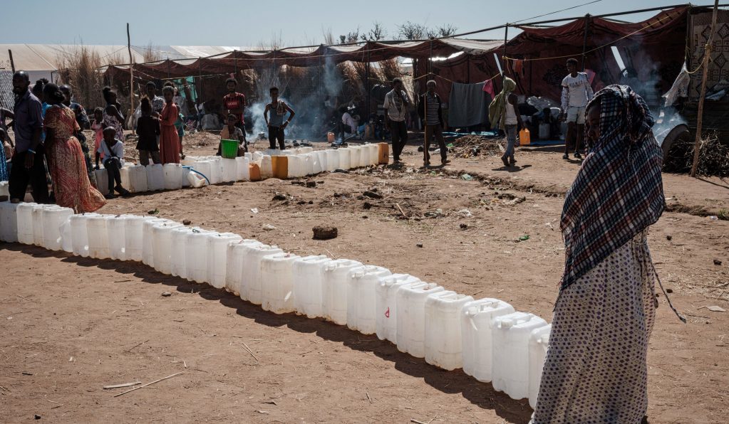 Ethiopian refugees who fled the Tigray conflict, wait to fill their jerrycans with water at Um Raquba reception camp in Sudan's eastern Gedaref state on December 3, 2020. The jerry cans are lined in an “L” shape and people cluster in groups talking.