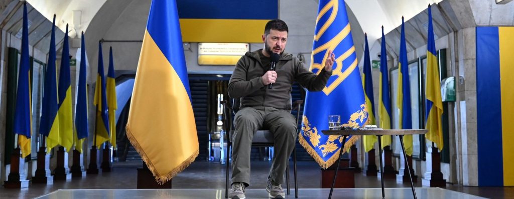 Ukrainian President Volodymyr Zelensky, sitting before Ukrainian national flags, addresses a press conference with international media in an underground metro station in Kyiv.