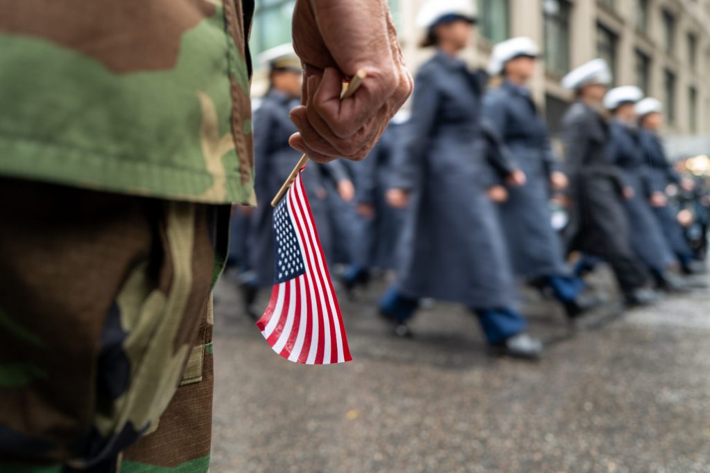Hand of a person in military fatigues holding a small U.S. flag in foreground, in front of servicemembers marching in blue uniforms, white hats, long blue coats on a parade route during a rainy day.