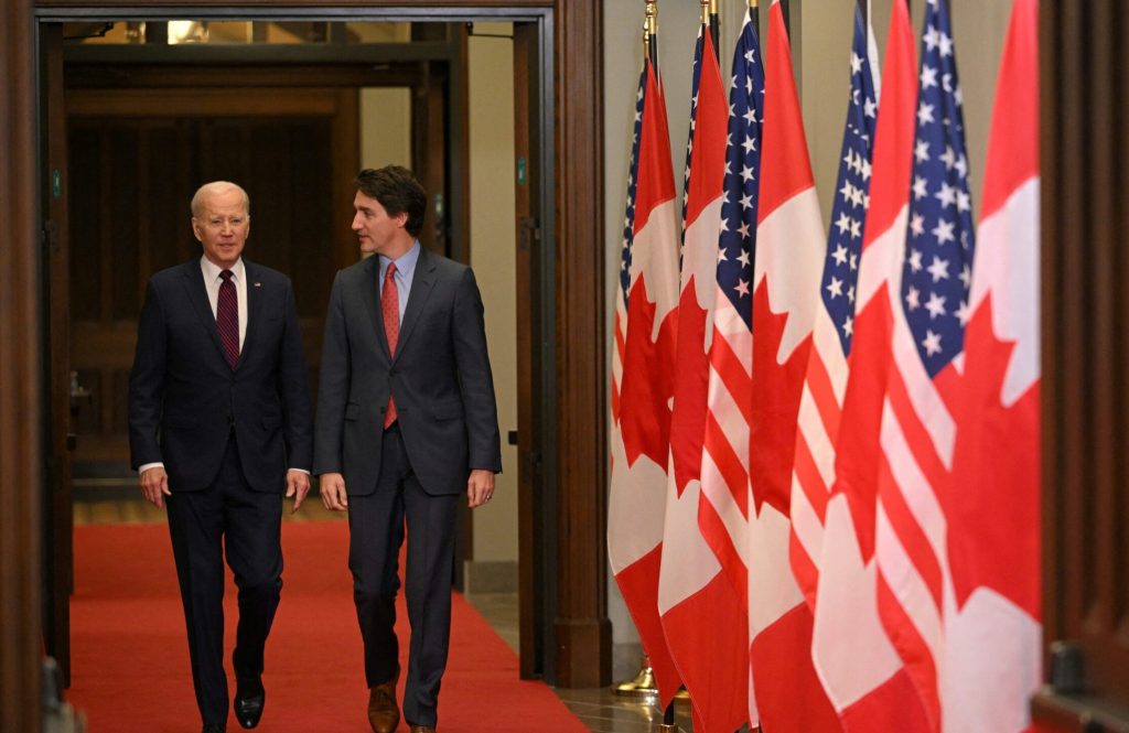 Biden and Trudeau walk together wearing dark suits and flanked by a line of U.S. and Canadian flags.