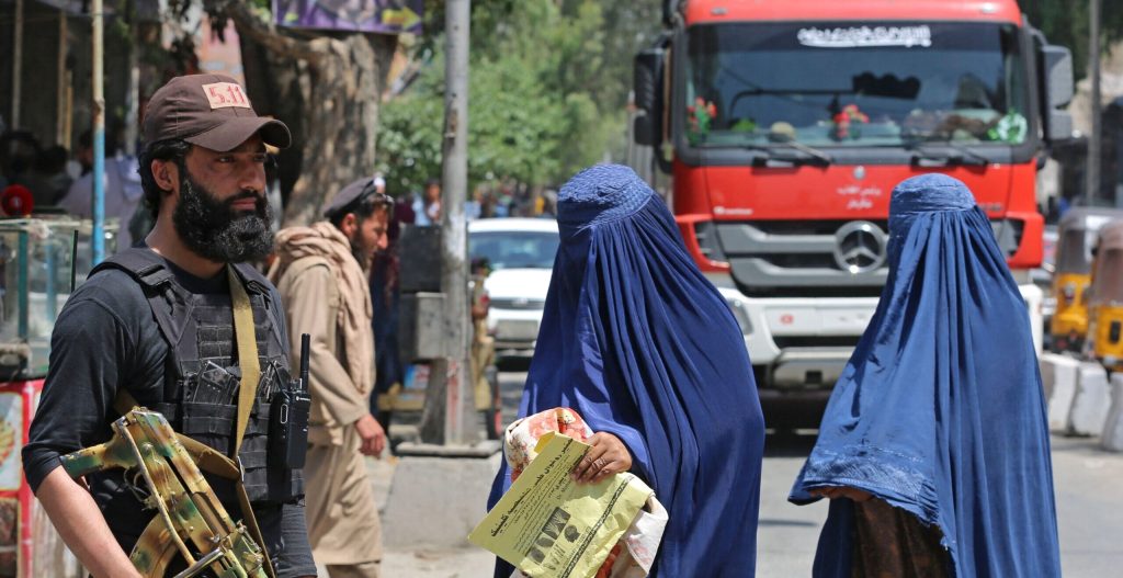 Afghan burqa-clad women walk past a Taliban security personnel along a street in Jalalabad
