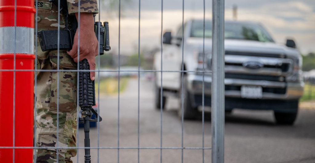 On the left, the lower body of a soldier holding a rifle behind a fence. One the right, a white truck.