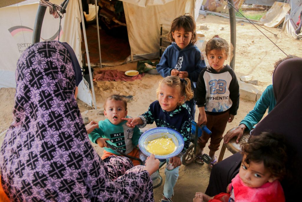 Four small children gather around as a woman offers them a bowl of yellow porridge.