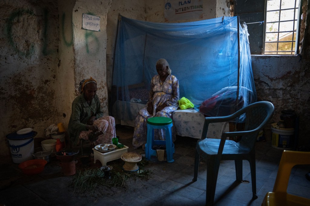 Two women sit inside a dark concrete shelter, one on a single bed draped in what appears to be blue mosquito netting, the other on a low stool, in Adwa, Ethiopia on March 30, 2025. A window with bars in the top right of the image provides a little light. Buckets and what appear to be cooking implements sit on the barren floor.