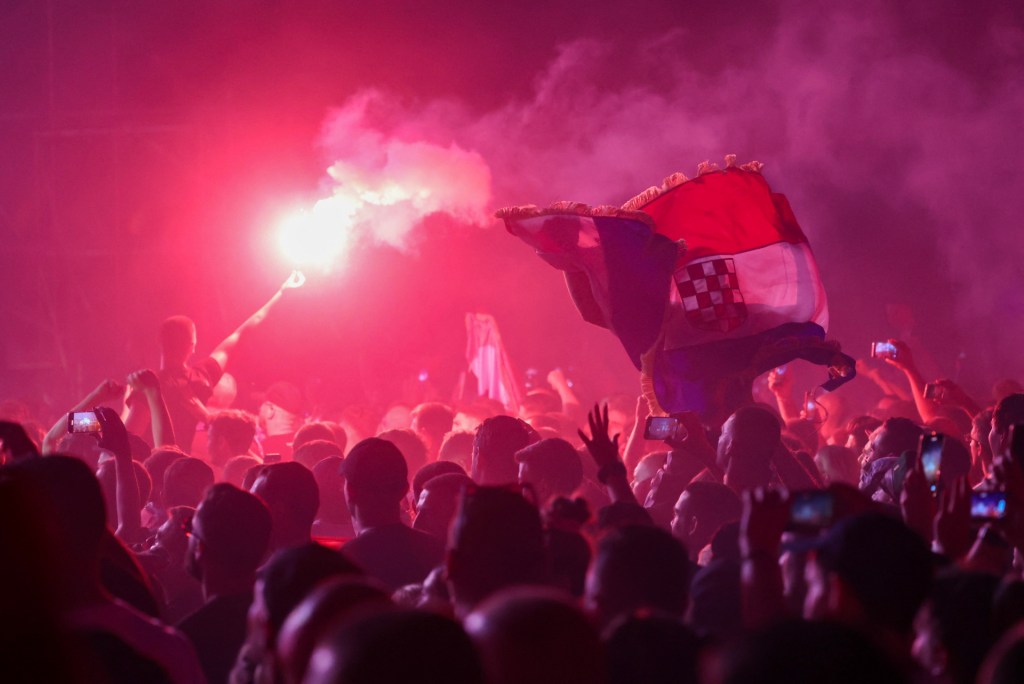 A Croatian national flag with its three bands of red, white and blue and a crest in the center, is seen weaving above fans at a concert, amid a smoke-filled, rose-colored backdrop and what look like small fireworks going off.