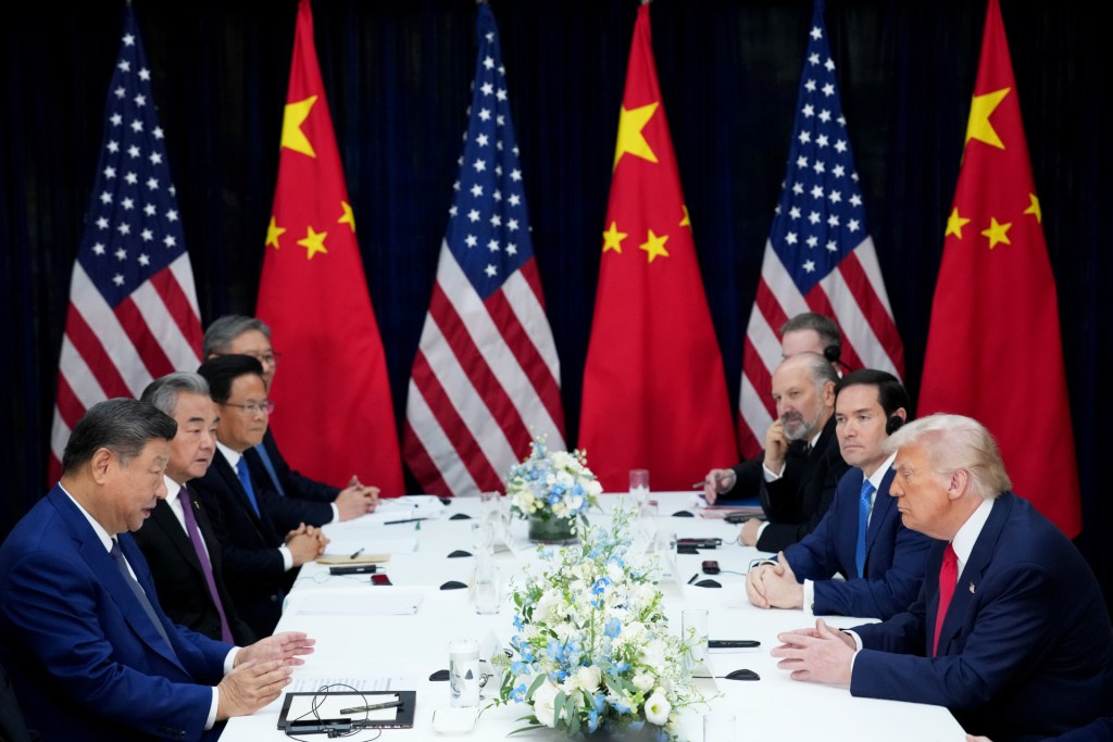 U.S. President Donald Trump (R) and Chinese President Xi Jinping (L) sit across from each other at a long table, each flanked on the right and left side of the image, respectively, by other officials, with three flags each of the United States and China, alternating along the back wall at the far end of the table.