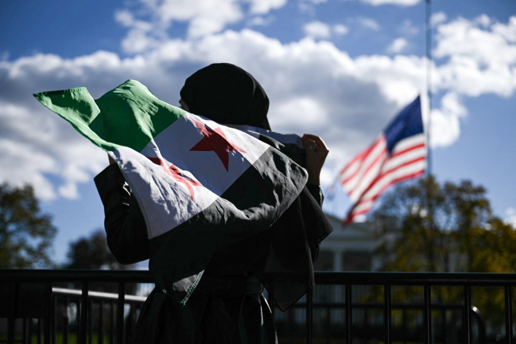 A man holds a Syrian flag across the street from the White House. An American flag waves in the background.