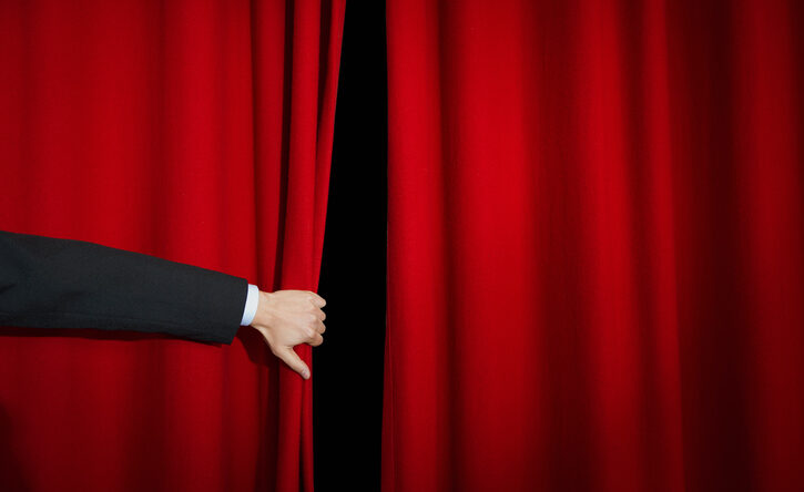 The hand of a person in a suit peels back a stage curtain.