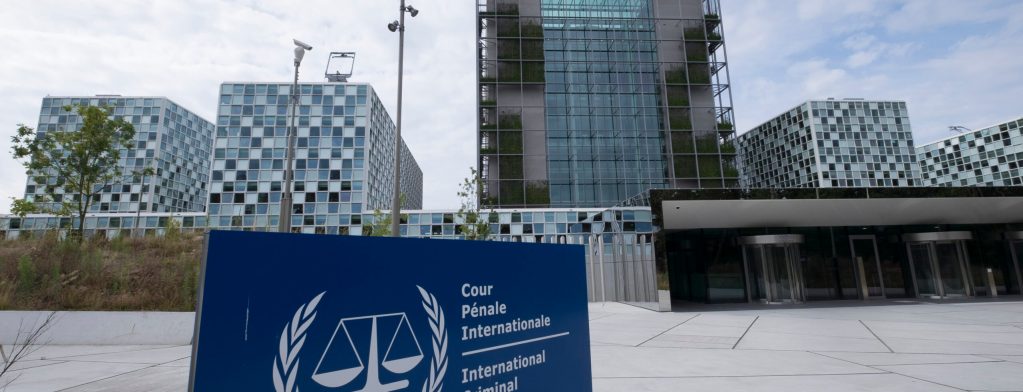 A sign with a dark blue background and white letters that reads "International Criminal Court" is seen with the building in the background.