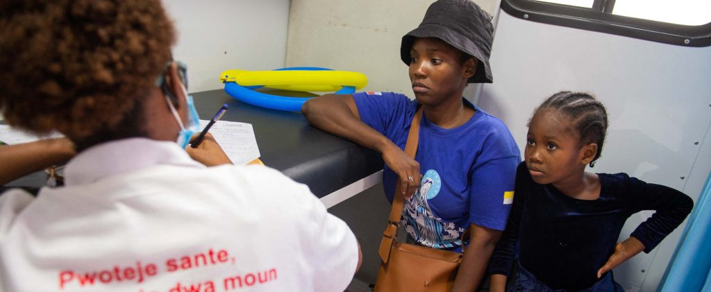 The photo shows the back of a health worker in the foreground wearing a shirt with donor logos on the back, speaking with the woman and her daughter in the background in a small room.