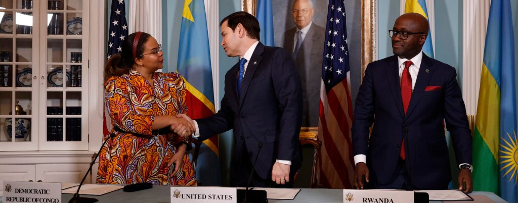 The three diplomats stand behind a desk where they have been signing the agreement. Behind them are a portrait on a blue wall and an array of two each of their three flags.