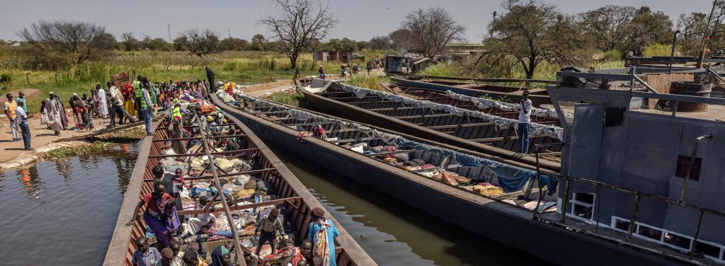The photo shows four very long, shallow docked boats arrayed next to each other diagonally across the image, seemingly made of iron or metal, carrying belongings and one with a number of adults and children in it. Other people are on land at the back of the image, next to a dirt road beside fields stretching into the distance.